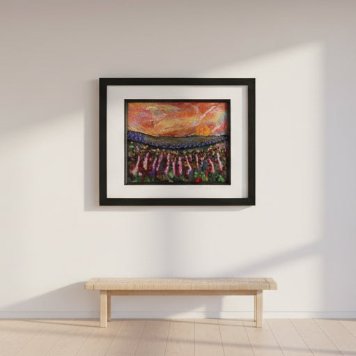 A framed art print depicting a colorful stadium scene with a dramatic sunset, displayed on a white wall above a light wood bench in a minimalist room setting.