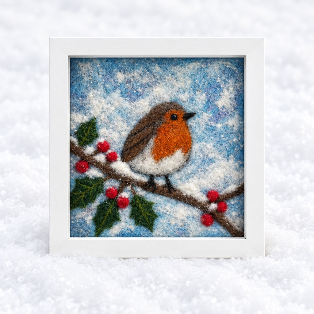 A square framed artwork showing a robin bird perched on a branch with red berries against a snowy blue background, displayed against a white backdrop with studio lights visible on the sides.