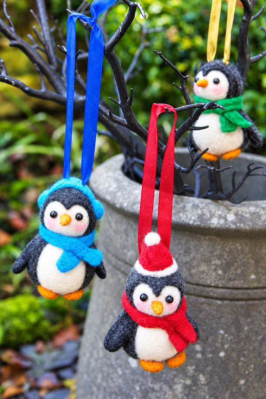 Three small felt penguin ornaments hang from bare black branches in a stone planter outdoors. One penguin wears a blue hat and scarf with a blue ribbon, one wears a red Santa hat and scarf with a red ribbon, and one wears a green scarf with a tan ribbon.