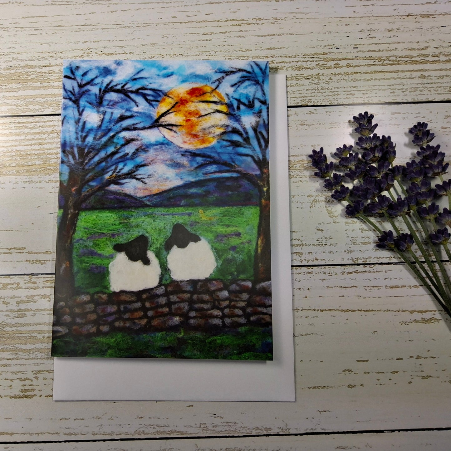 A square art print or card featuring a painted scene of two sheep on a stone wall in a green field under a full moon,