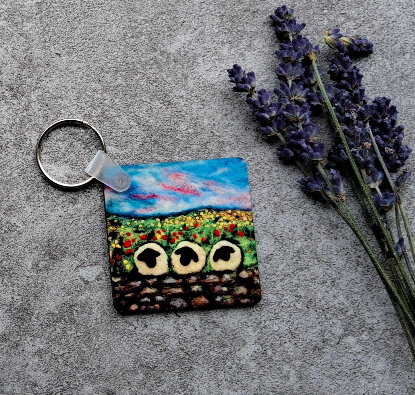 A small square keychain with a colorful landscape painting of sunflowers and a blue sky, attached to a silver metal ring, displayed on a gray concrete surface next to lavender sprigs.