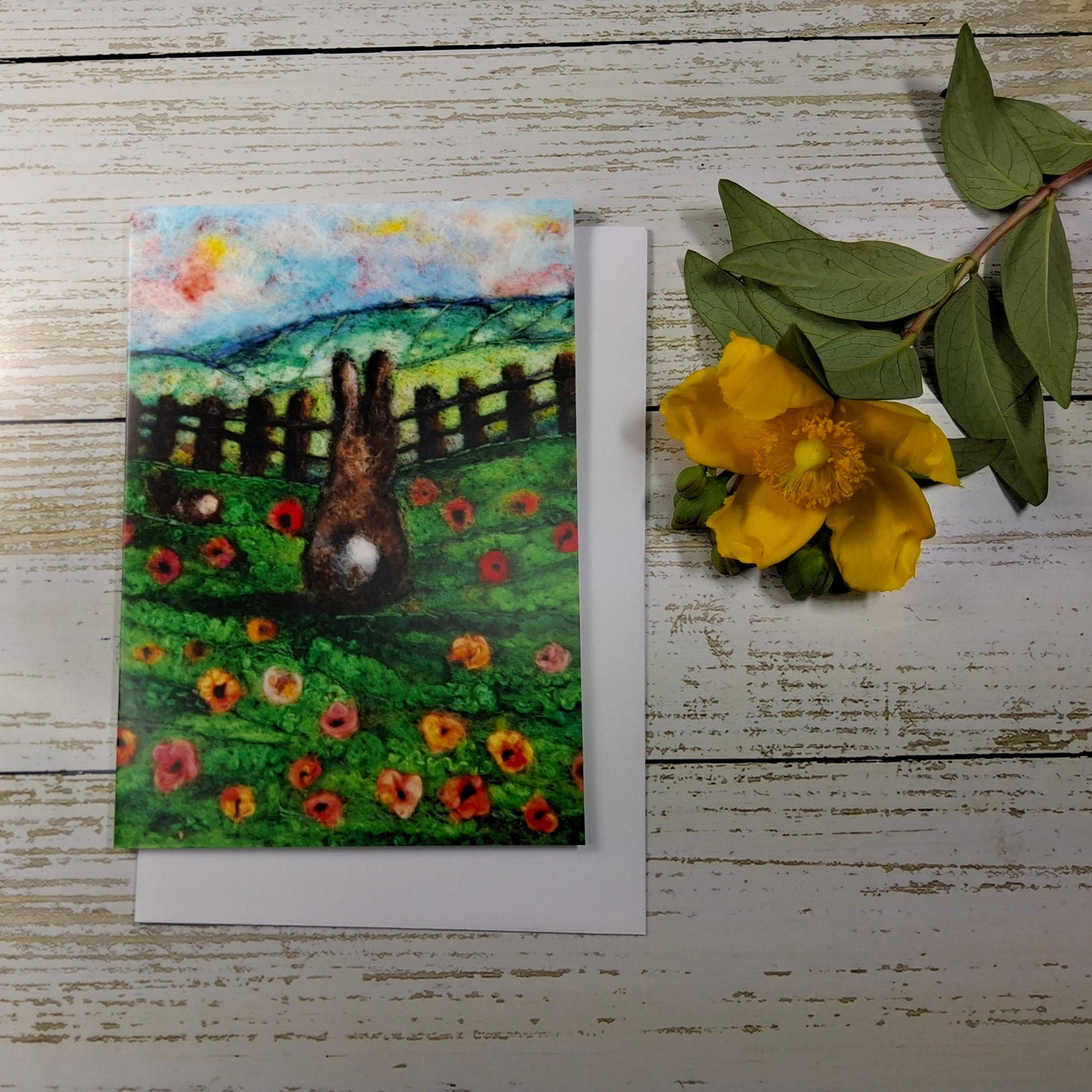 A square art print or card featuring a painted scene of a rabbit in a flower field with a fence and hills, placed on a white surface next to a yellow flower and green leaves.