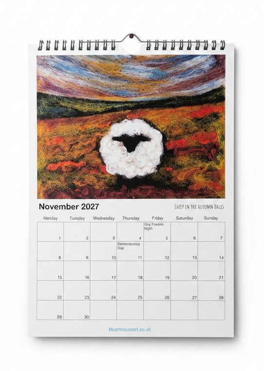 A spiral-bound November 2027 wall calendar page featuring a colorful painted scene of a sheep in a field, photographed against a light wood background.