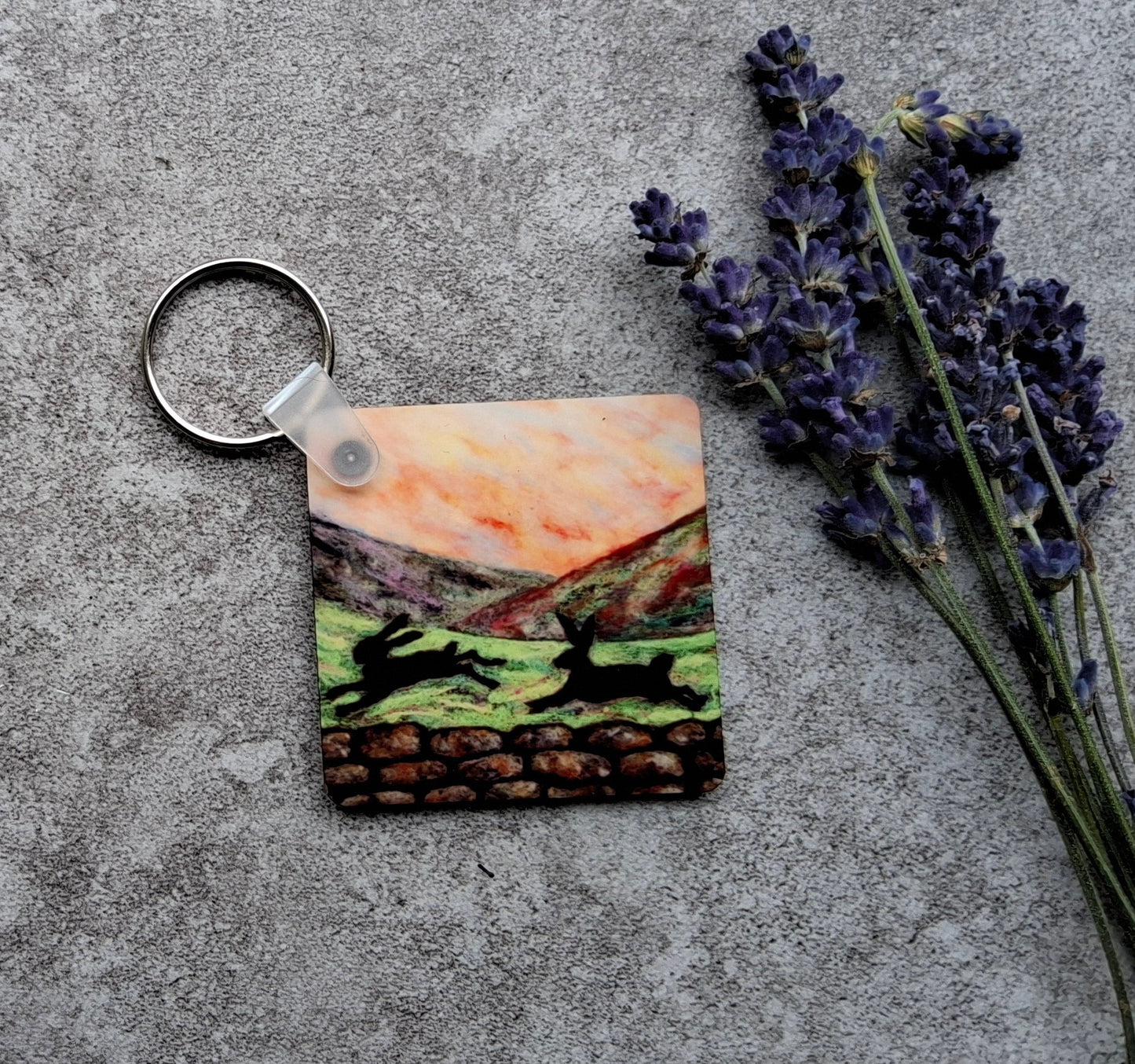 A square acrylic keychain featuring a colorful landscape illustration of deer in a field, resting on a textured gray concrete surface next to dried lavender sprigs.