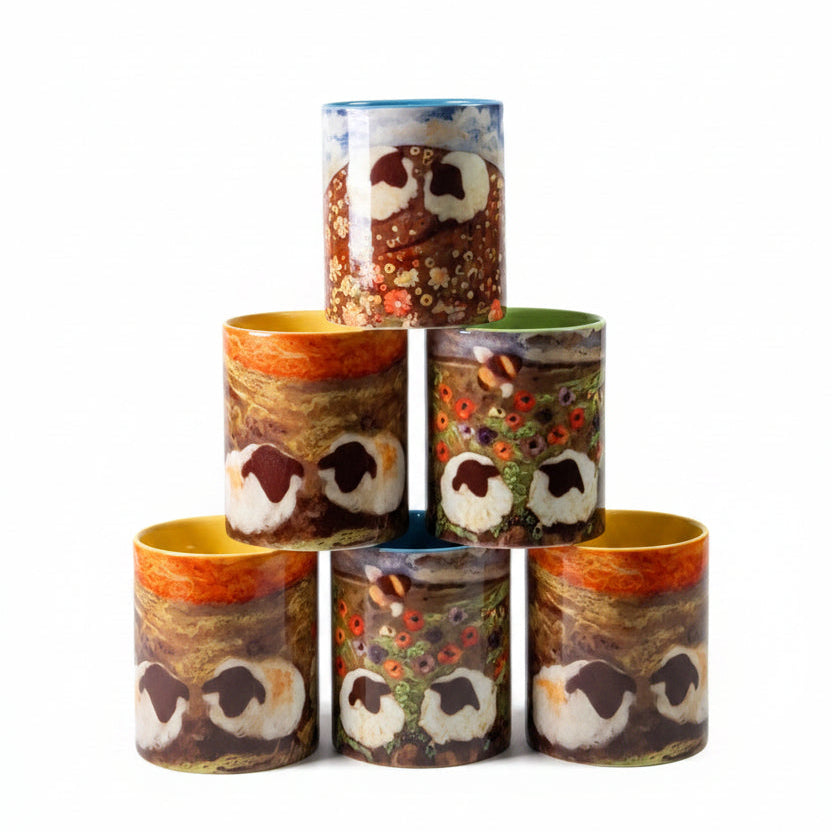 Four owl-themed cylindrical cups stacked in a pyramid on a light wood tabletop, with a large textured panel behind them and a desk lamp visible in the background.
