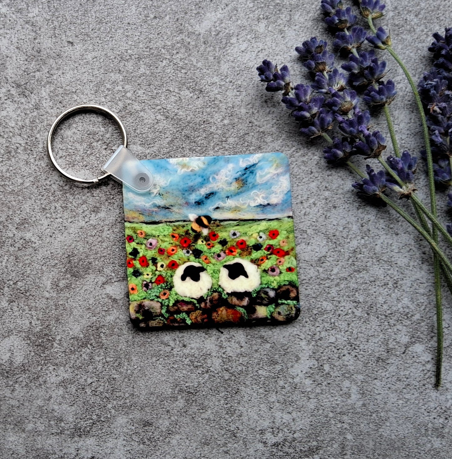 A square keychain featuring a colorful painted landscape with cows in a field under a blue sky, displayed on a textured gray surface next to sprigs of lavender.