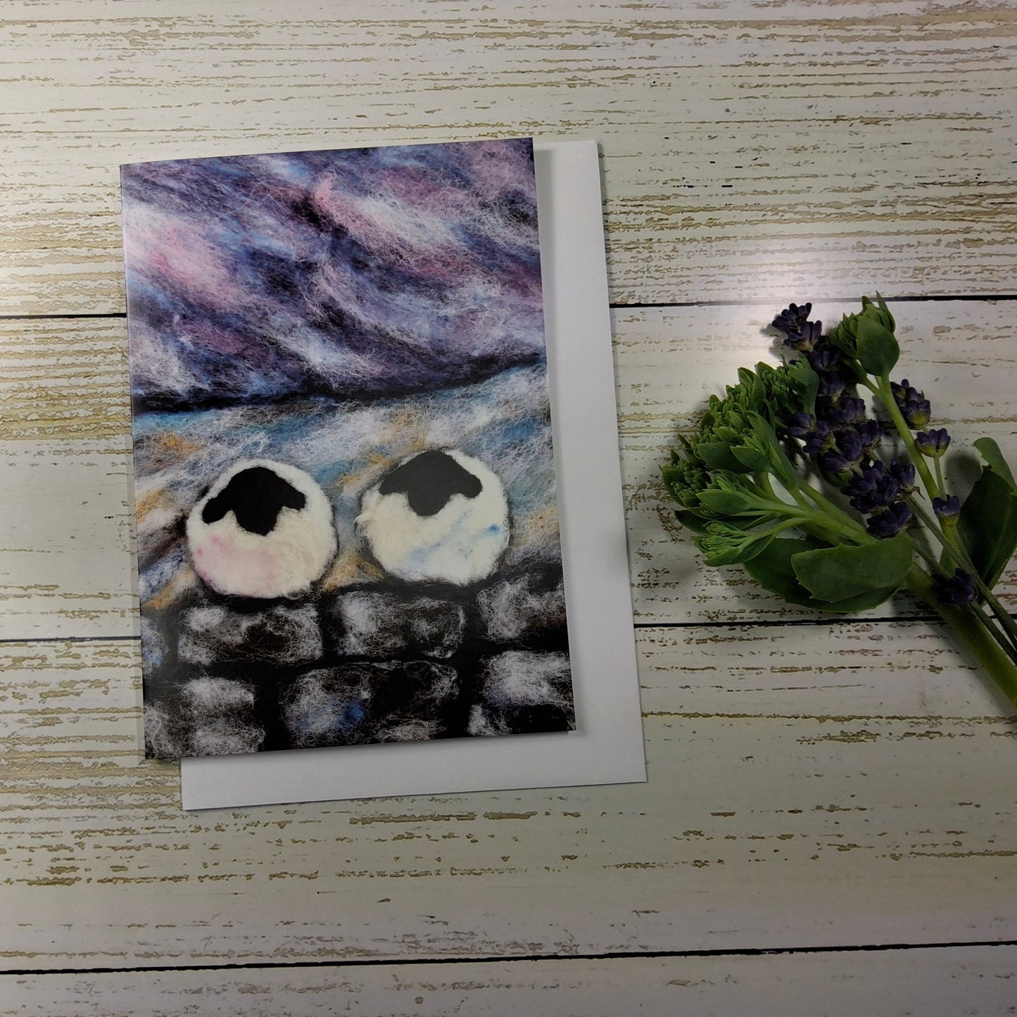 A square art print or card featuring a painted scene of two white birds with black heads perched on a dark stone wall under a purple and blue sky, displayed on a whitewashed wooden surface next to a small bouquet of purple flowers.