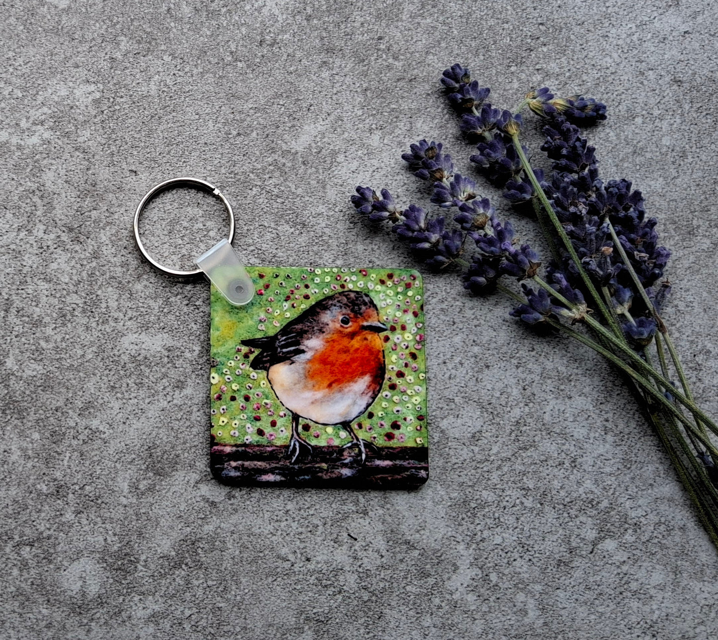 A square keychain featuring a painted robin bird on a green textured background, attached to a silver split ring and placed on a gray concrete surface next to dried lavender sprigs.