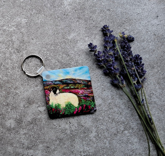 A square painted keychain featuring a white sheep in a colorful landscape, attached to a silver metal ring, styled on a textured gray concrete surface with dried lavender and yellow flowers.