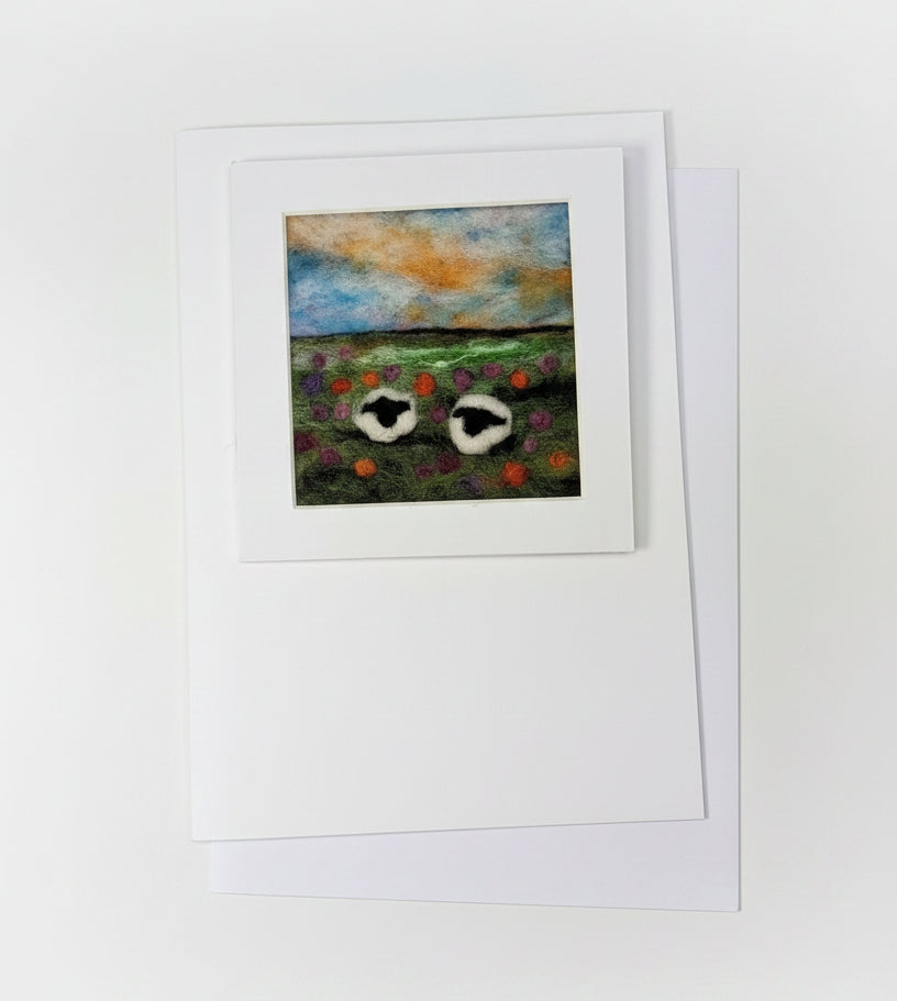 A small framed art print featuring two white birds with red accents against a blue and orange sky, displayed on a white card and envelope over a rustic whitewashed wooden surface with decorative greenery.
