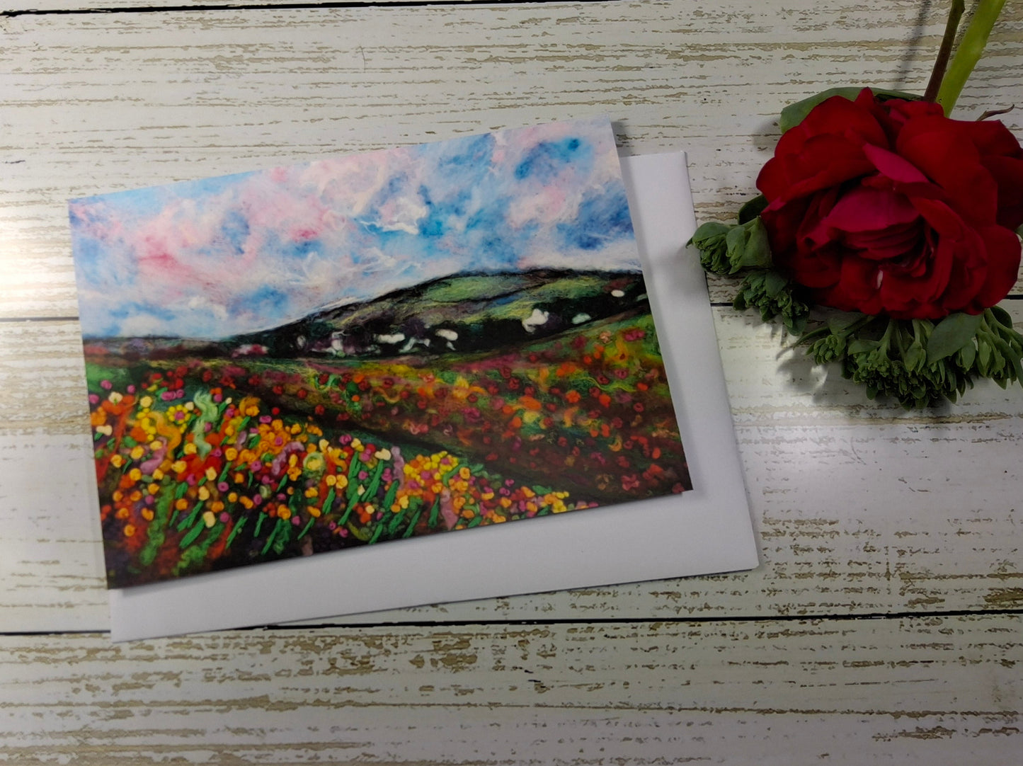 A square landscape painting of a flower field under a blue and pink sky, displayed on a white wooden surface next to a red rose.