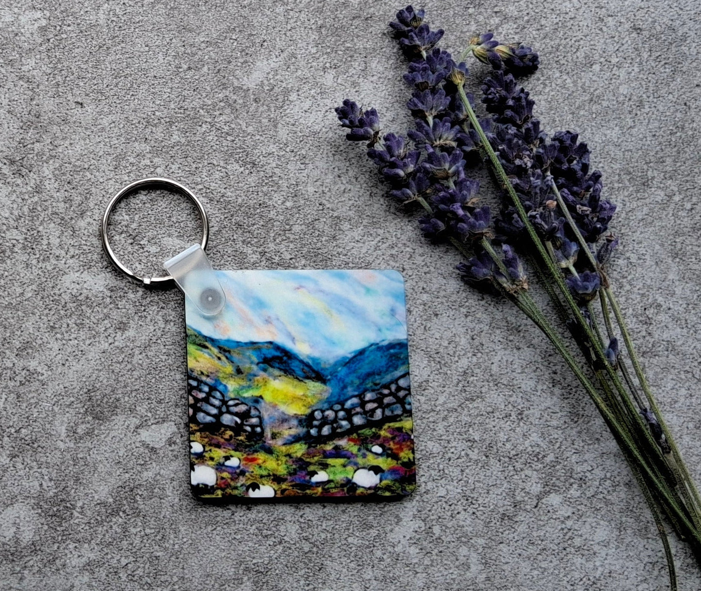 A small square keychain featuring a colorful landscape painting of mountains and lavender fields, displayed on a gray concrete surface next to dried lavender sprigs.