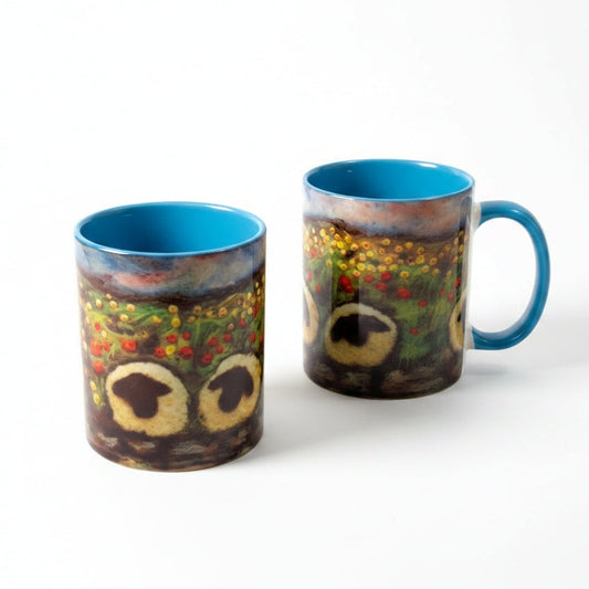 Two ceramic mugs with a colorful owl face design and bright blue interiors, placed on a white wooden tabletop next to a small sprig of greenery.