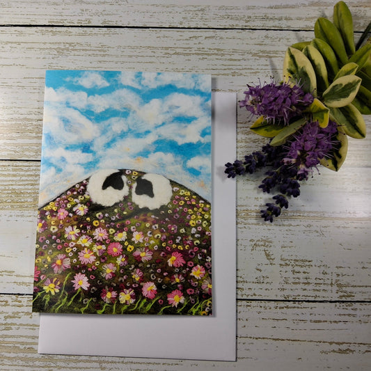 A square art print or card featuring a whimsical landscape with two black-and-white sheep faces above a field of colorful wildflowers under a blue sky, displayed on a white envelope over a whitewashed wood surface with decorative flowers nearby.