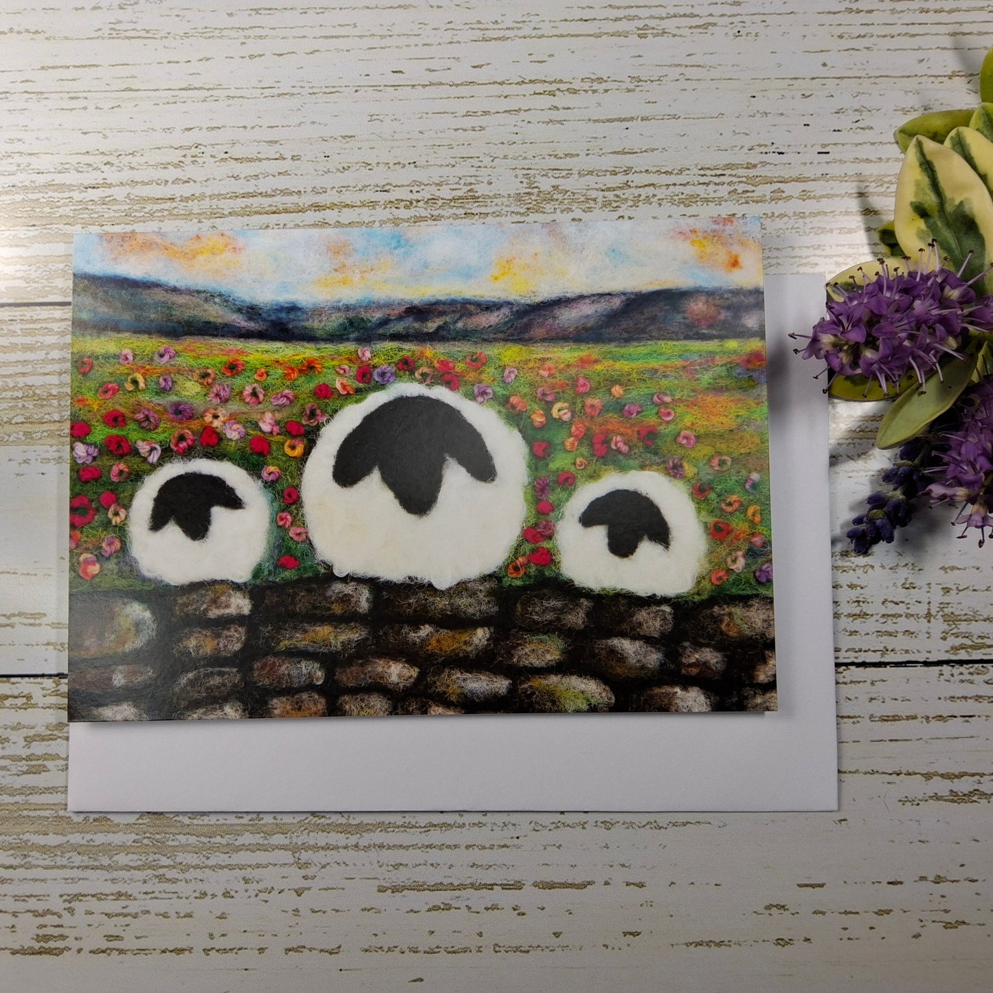 A square art print or card featuring a painted pastoral landscape with three white sheep in front of a stone wall, displayed on a whitewashed wood surface next to green leaves and purple flowers.