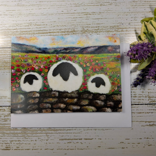 A square art print or card featuring a painted pastoral landscape with three white sheep in front of a stone wall, displayed on a whitewashed wood surface next to green leaves and purple flowers.