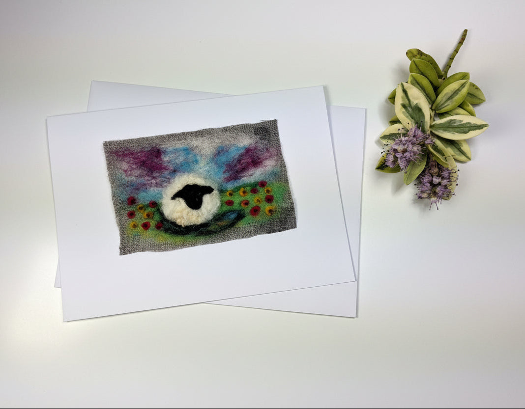 A small handmade felt painting of a sheep on a textured white card, displayed on a rustic white wooden table next to a small plant sprig.