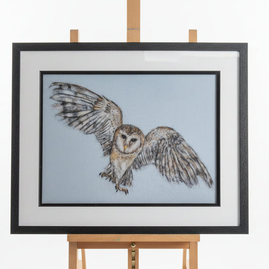 A framed owl artwork displayed on an easel in a workshop. The piece shows a detailed owl with wings spread against a pale blue-gray background, enclosed in a black frame with a wide white mat.