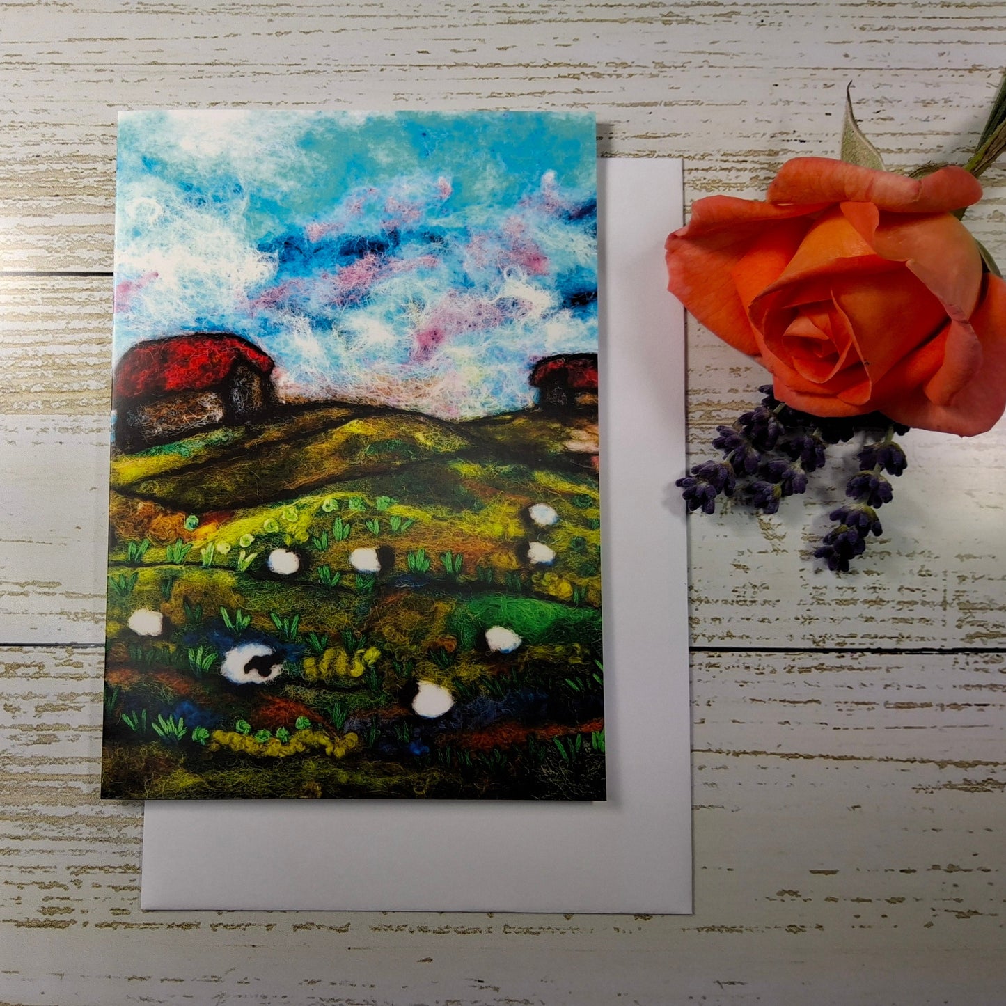 A square landscape art print or card featuring an impressionistic painted field with green hills, white sheep, red barns, and a blue sky,