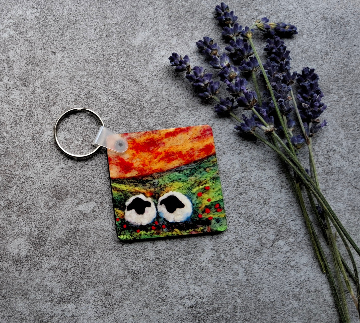 A square keychain with a colorful, abstract painted face design in orange, green, and red, attached to a silver metal key ring. It is styled on a textured gray concrete surface next to sprigs of dried lavender.