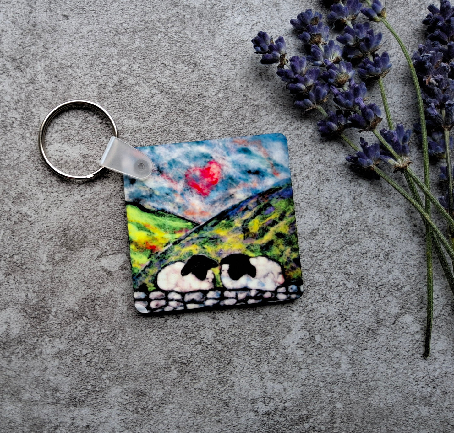 A square keychain featuring a colorful painted landscape with sheep, set on a textured gray surface next to sprigs of lavender.