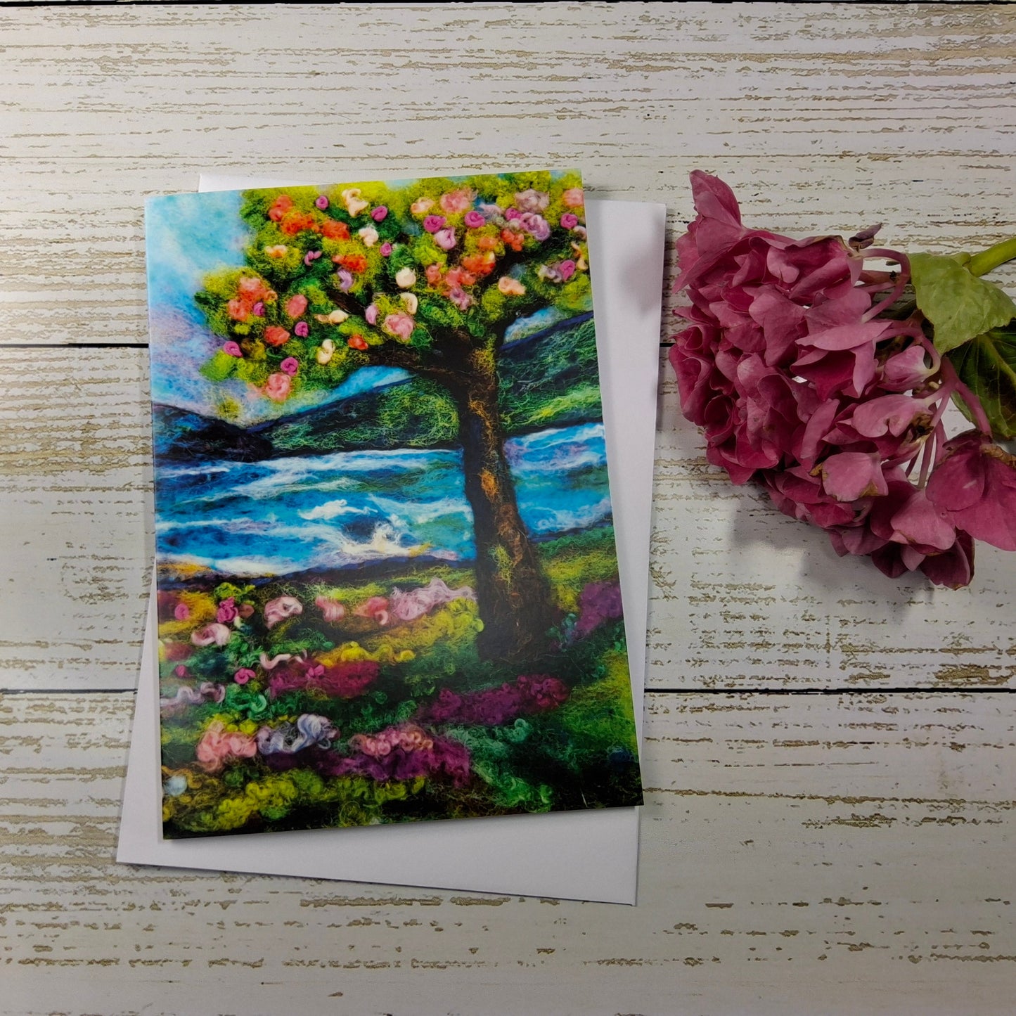 A square art print or card featuring an expressive painting of a flowering tree by a blue water landscape, styled on a white wood background next to a pink hydrangea flower.