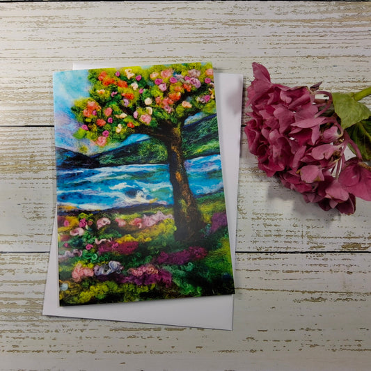 A square art print or card featuring an expressive painting of a flowering tree by a blue water landscape, styled on a white wood background next to a pink hydrangea flower.