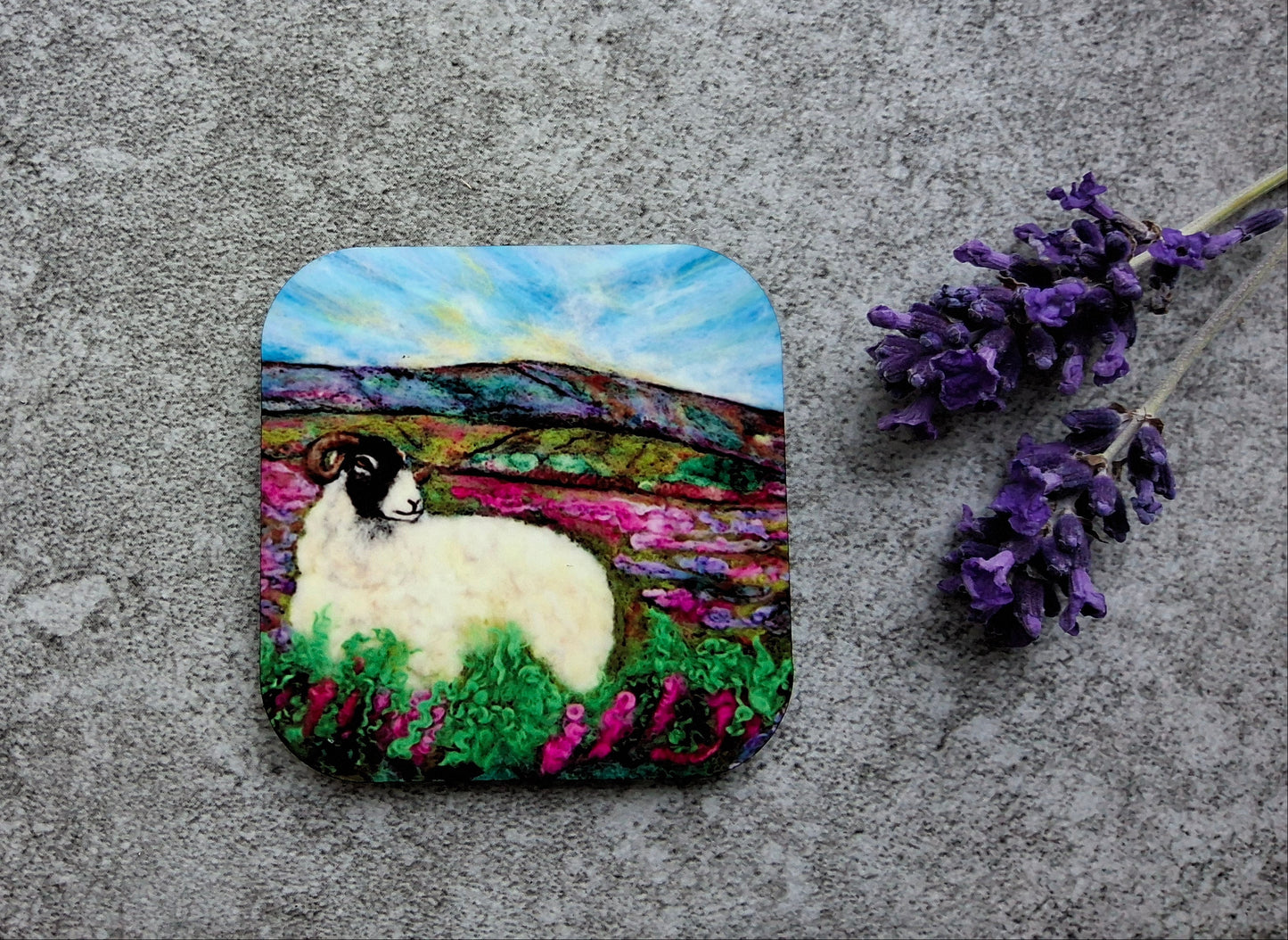 A square coaster featuring a colorful painted scene of a white sheep with a black head in a field of pink flowers under a blue sky, displayed on a textured gray surface next to purple lavender sprigs.