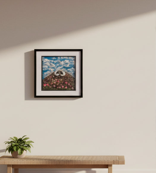 A framed square art print hanging on a beige wall, featuring a whimsical barn scene with a panda on the roof under a blue cloudy sky, next to another framed print.