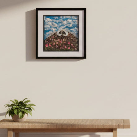 A framed square art print hanging on a beige wall, featuring a whimsical barn scene with a panda on the roof under a blue cloudy sky, next to another framed print.