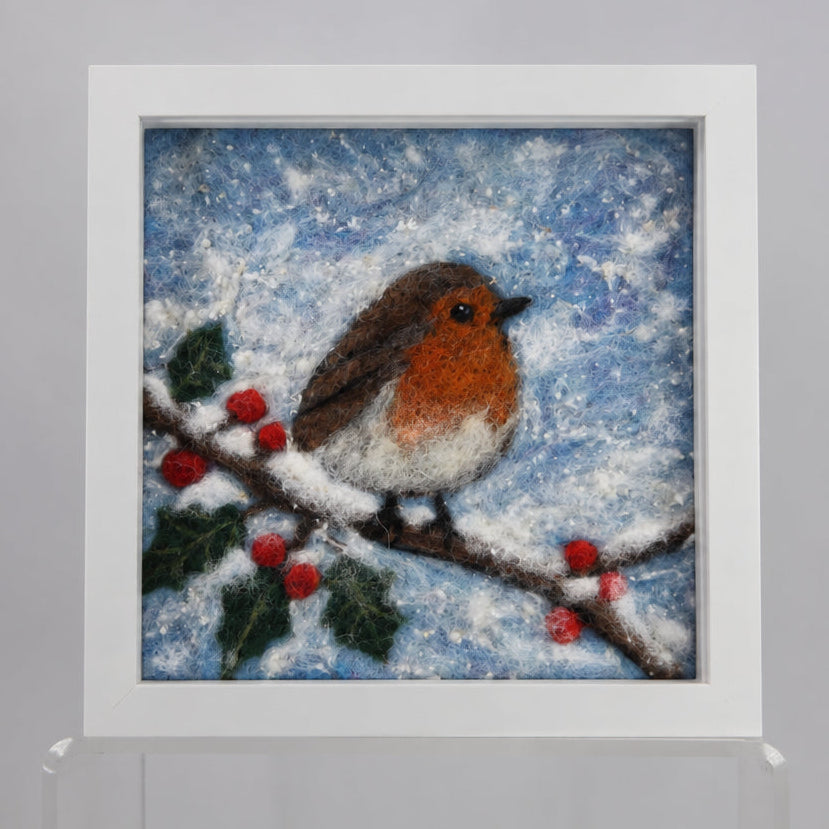 A framed needlepoint artwork featuring a robin bird perched on a branch with red berries against a snowy blue background, displayed on a white wall.