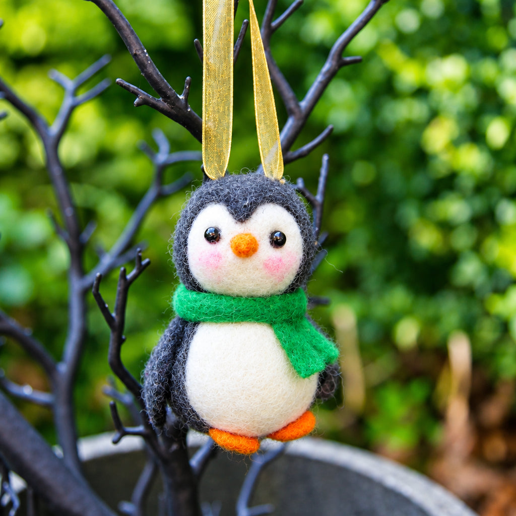 A small felted penguin ornament with a green scarf and orange beak feet hangs from a gold ribbon against a dark decorative branch and blurred garden background.