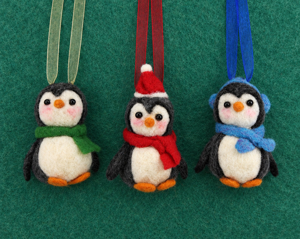 Three felt penguin ornaments hanging from ribbons against a green felt background. One penguin wears a green scarf, one wears a red Santa hat and red scarf, and one wears a blue scarf.