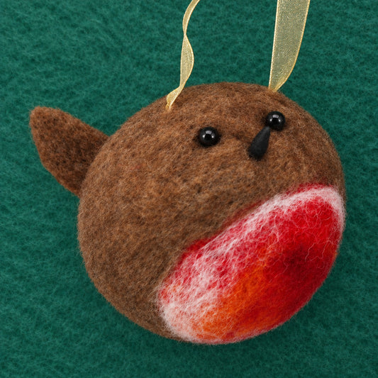 A handmade felted wool bird ornament, likely a robin, with brown feathers, a red and orange beak, black bead eyes, and a gold ribbon hanger, set against a textured green felt background.