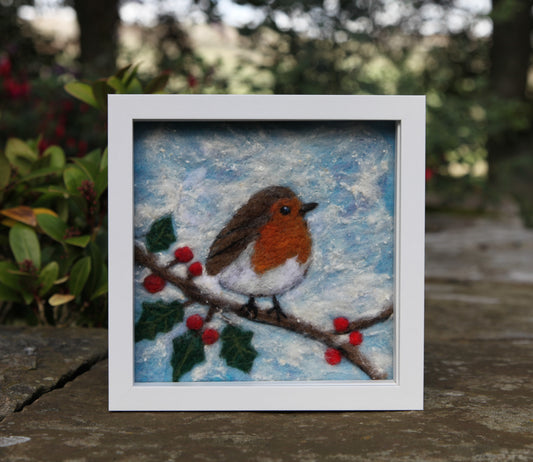 A framed needlepoint artwork featuring a robin bird perched on a branch with red berries against a snowy blue background, displayed outdoors on a stone surface.