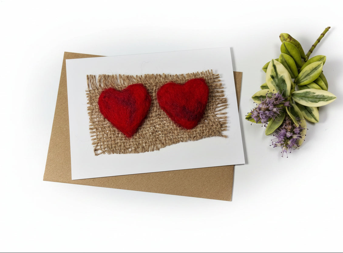 A handmade greeting card featuring two red felt hearts on a burlap background, placed on top of a brown kraft envelope on a distressed white wood surface with decorative greenery in the corner.