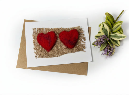 A handmade greeting card featuring two red felt hearts on a burlap background, placed on top of a brown kraft envelope on a distressed white wood surface with decorative greenery in the corner.