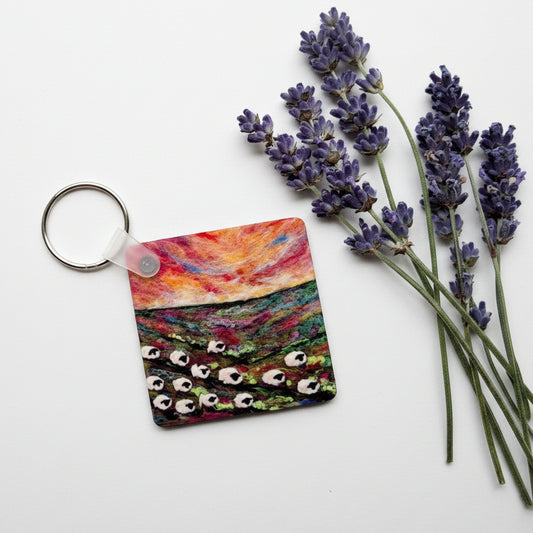 A square leather keychain with a colorful landscape painting print and a silver split ring, styled on a gray concrete surface next to dried lavender sprigs.