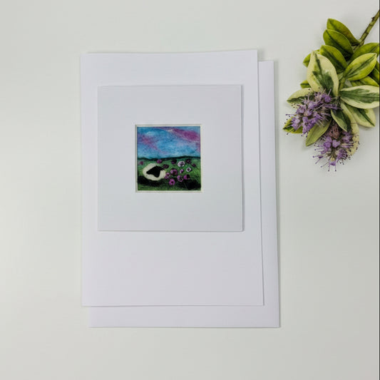 A square art print with a wide white mat is displayed on a distressed white wood tabletop, alongside a small dried floral sprig. The print features a colorful landscape painting with a blue and purple sky, green foreground, and a dark boat.
