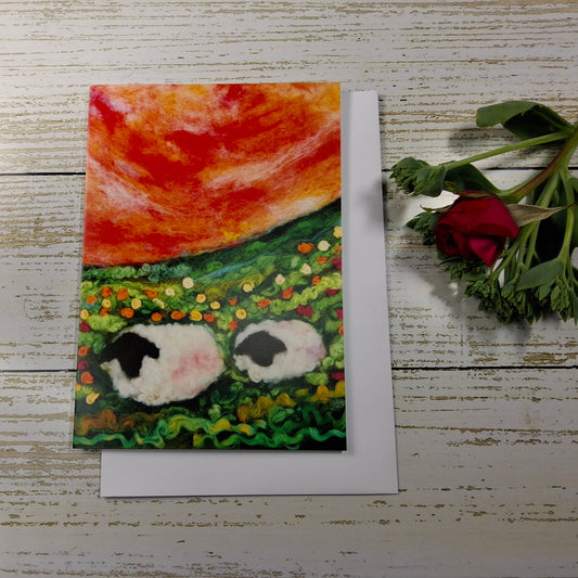 A square greeting card featuring a colorful painting of two sheep in a field under a bright orange sky, placed on a white wooden surface next to a red rose stem.