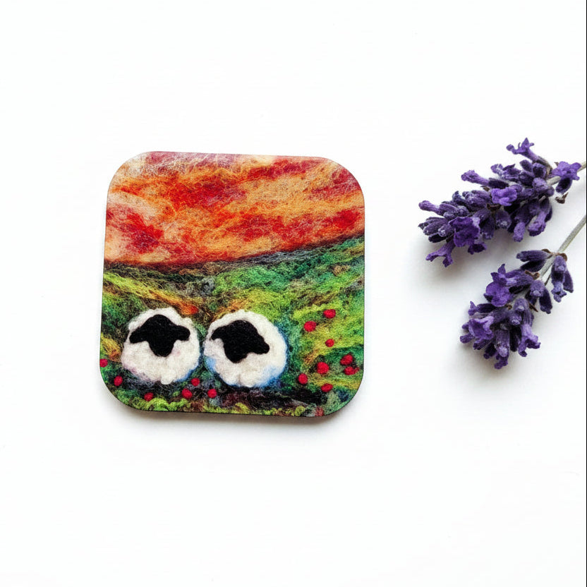 A square ceramic coaster with rounded corners featuring a painted landscape of a red and orange sunset over a green field with sheep, displayed on a gray textured surface next to a sprig of purple lavender.