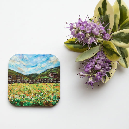 A square coaster featuring a painted landscape of a green field under a blue sky, styled on a gray textured surface next to purple flowers.