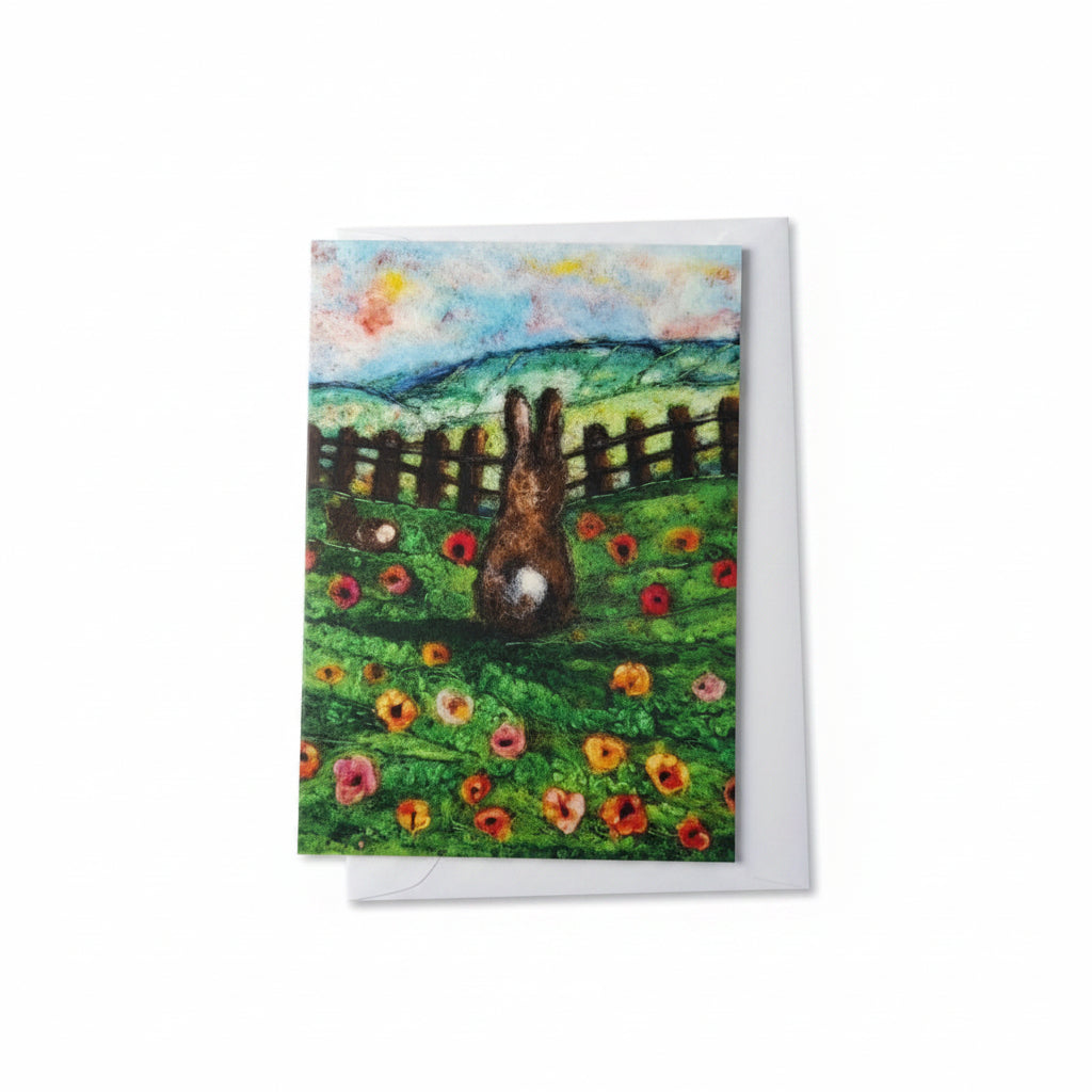 A square art print or card featuring a painted scene of a rabbit in a flower field with a fence and blue sky, placed on a white wooden surface next to a yellow flower.