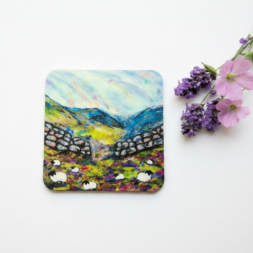 A square coaster featuring a colorful painted landscape of sheep in a field with stone walls and blue mountains under a cloudy sky, styled on a white surface next to pink and purple flowers.