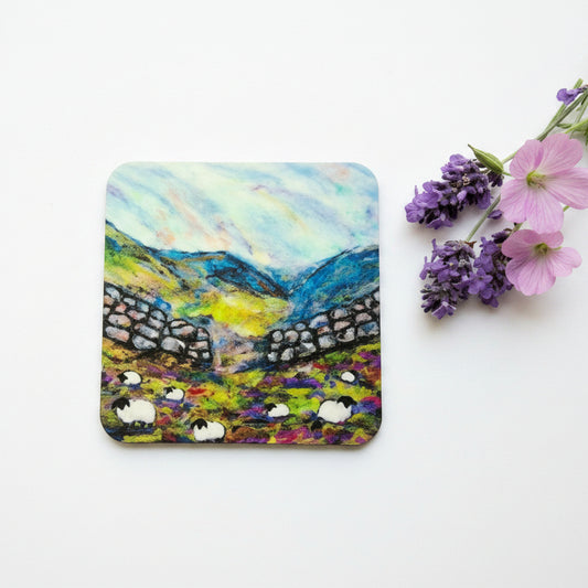 A square coaster featuring a colorful painted landscape of sheep in a field with stone walls and blue mountains under a cloudy sky, styled on a white surface next to pink and purple flowers.