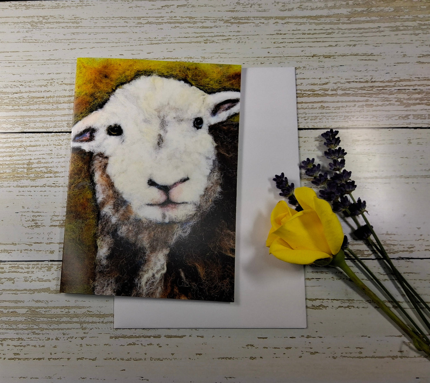 A square art print featuring a painted white sheep face with a yellow and brown textured background, displayed on a white sheet of paper over a distressed white wood surface, styled with a yellow flower and lavender sprigs.