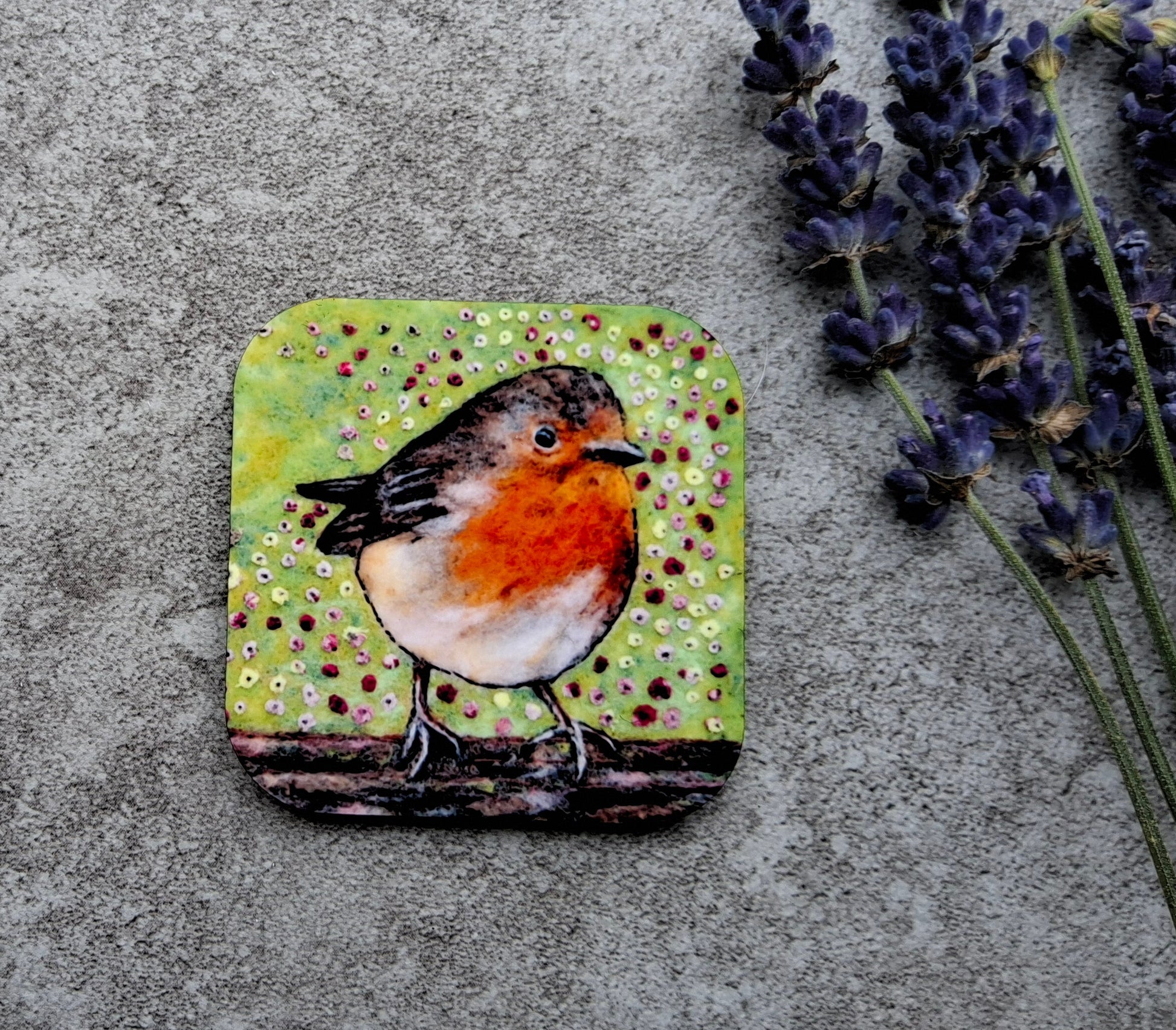 A square coaster featuring a painted robin bird on a green spotted background, styled on a textured gray surface next to lavender sprigs.