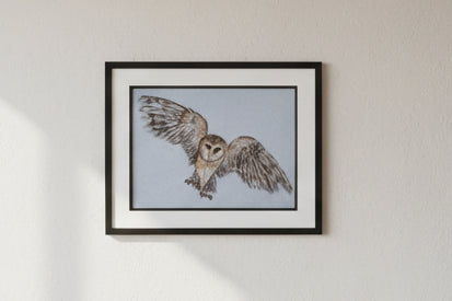 A framed owl artwork displayed on a wooden easel in a cluttered craft or studio room. The artwork shows a detailed owl with spread wings in brown, gray, and beige tones on a light background, enclosed in a black frame with a wide white mat.