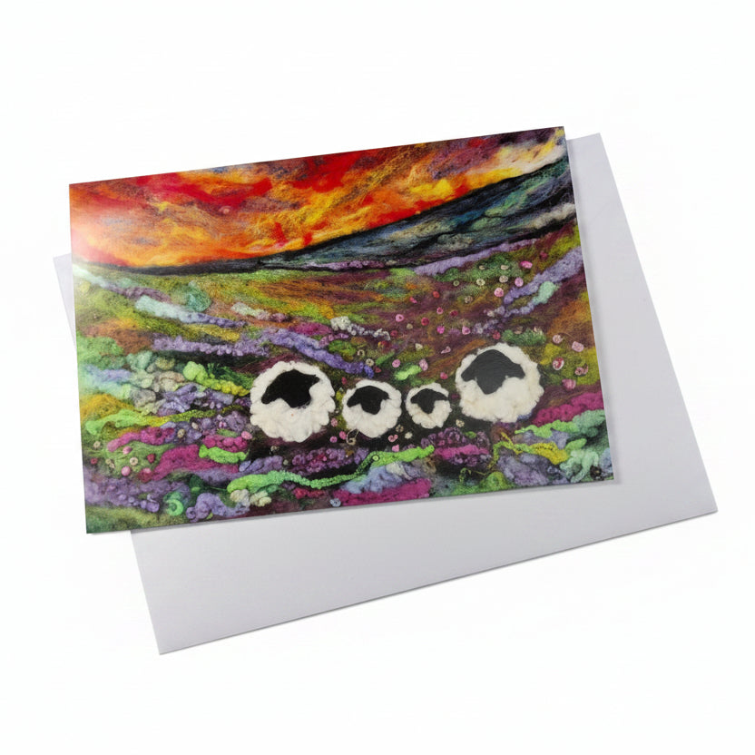 A square art print or greeting card with a colorful landscape painting of three sheep, placed on a white envelope 