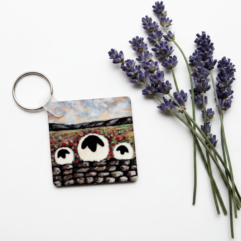 A square acrylic keychain featuring a hand-painted scene of sheep in a field, placed on a textured gray surface next to sprigs of lavender.